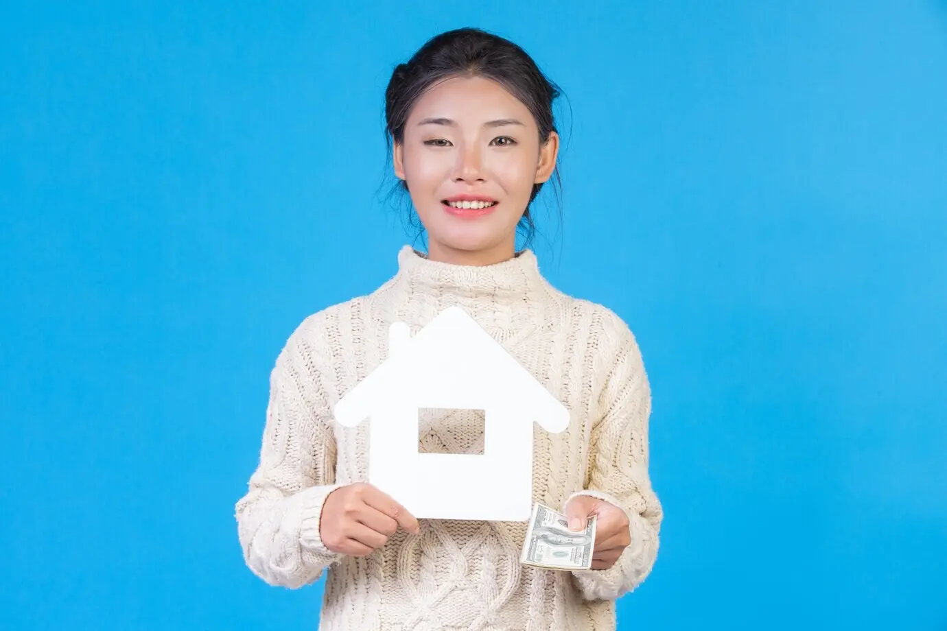 Eine schöne Frau, die einen neuen, langärmeligen, weißen Teppich trägt, hält das Haussymbol und Dollarscheine vor blauem Hintergrund. Handel.